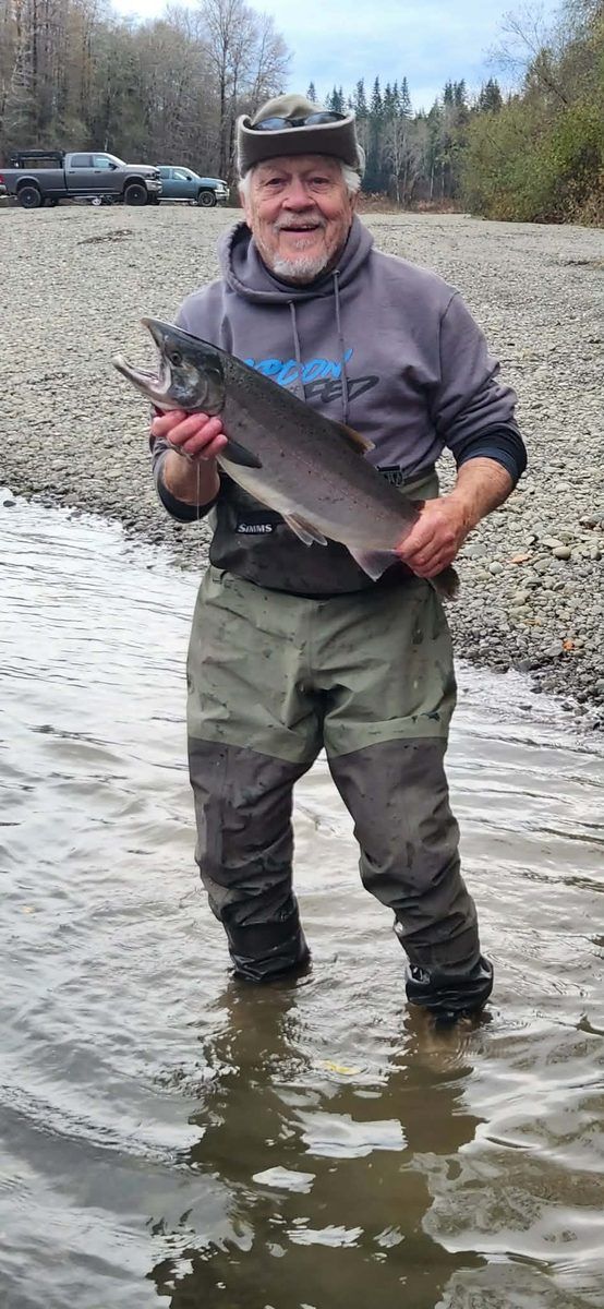 An angler standing on a gravel bar holding a large steelhead, pickup trucks parked in the distance