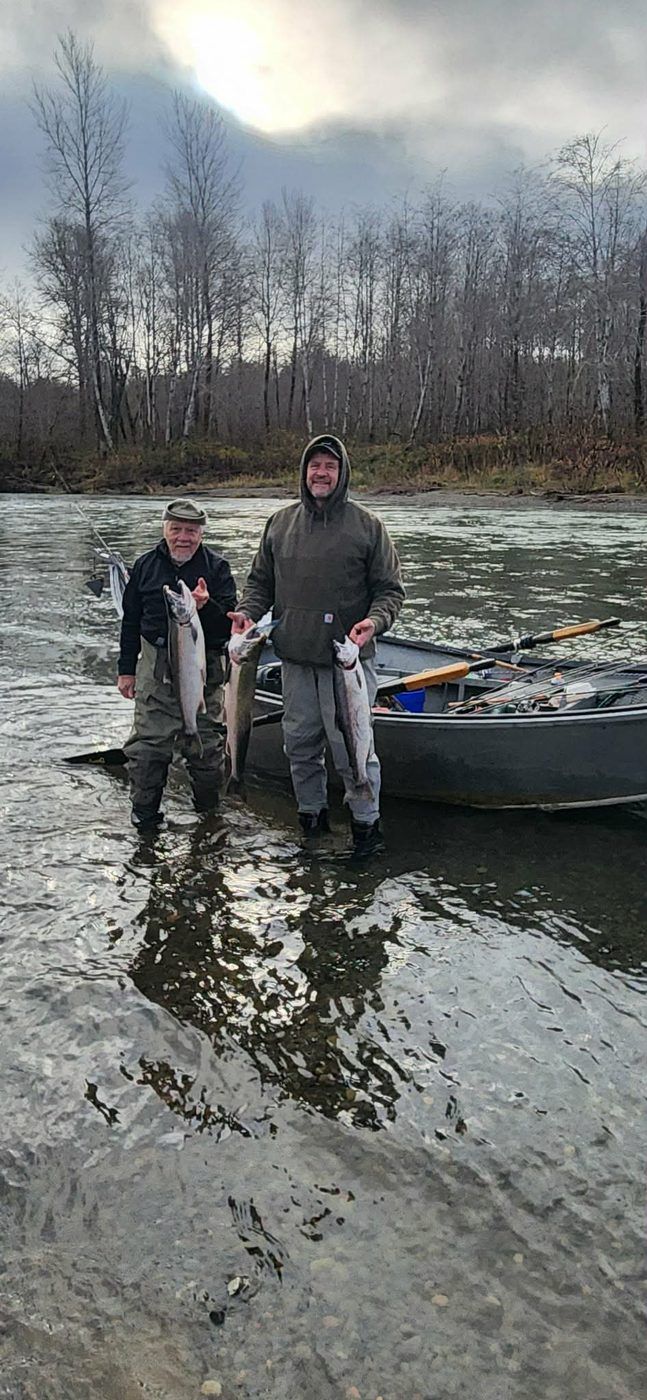 Two anglers standing in shallow water beside the drift boat, each holding salmon, with a forested riverbank behind